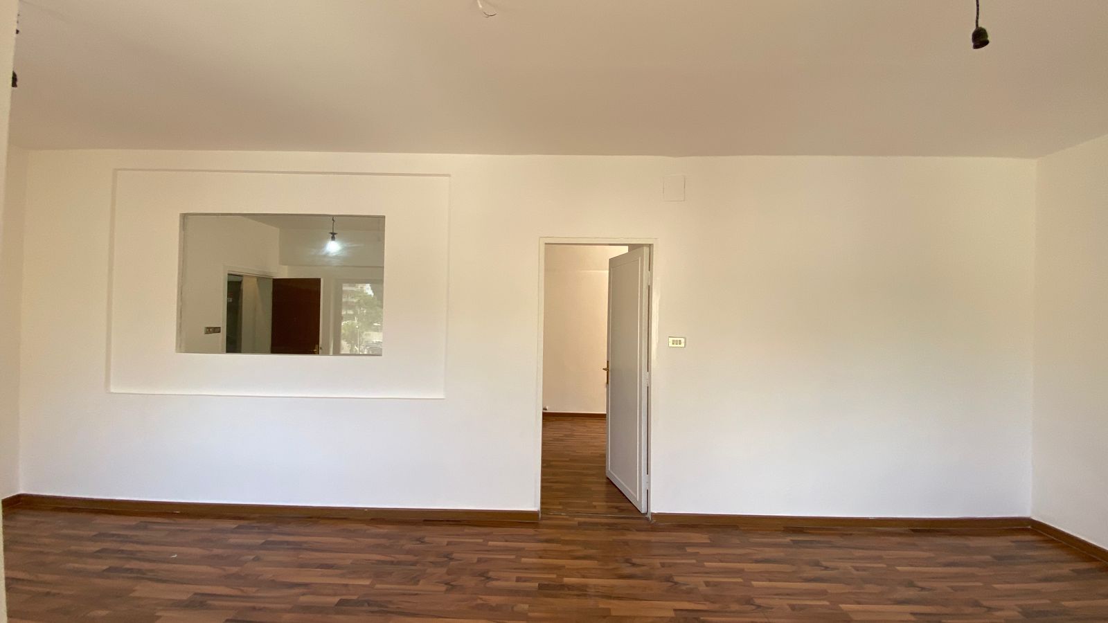 b79e9da4-f383-4e2a-b556-096d679c306f Empty white-walled room with dark wood floor; an open doorway leads to another space, and a large wall window reveals a glimpse of a next room.