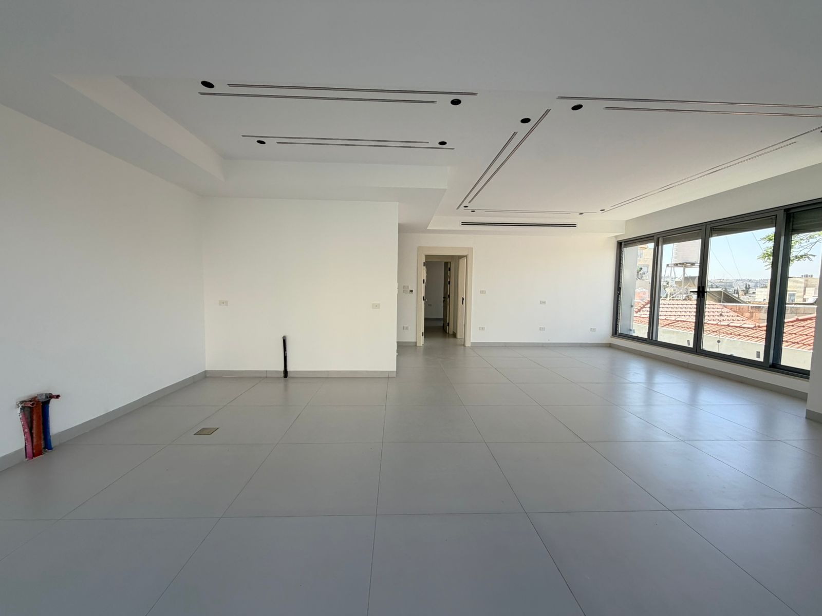 b7ecc09e-5e3a-4714-83a4-ef88d93749a4 Bright empty open-plan room with large windows, white walls and light-gray tiled floor; doorway center and recessed ceiling details visible.