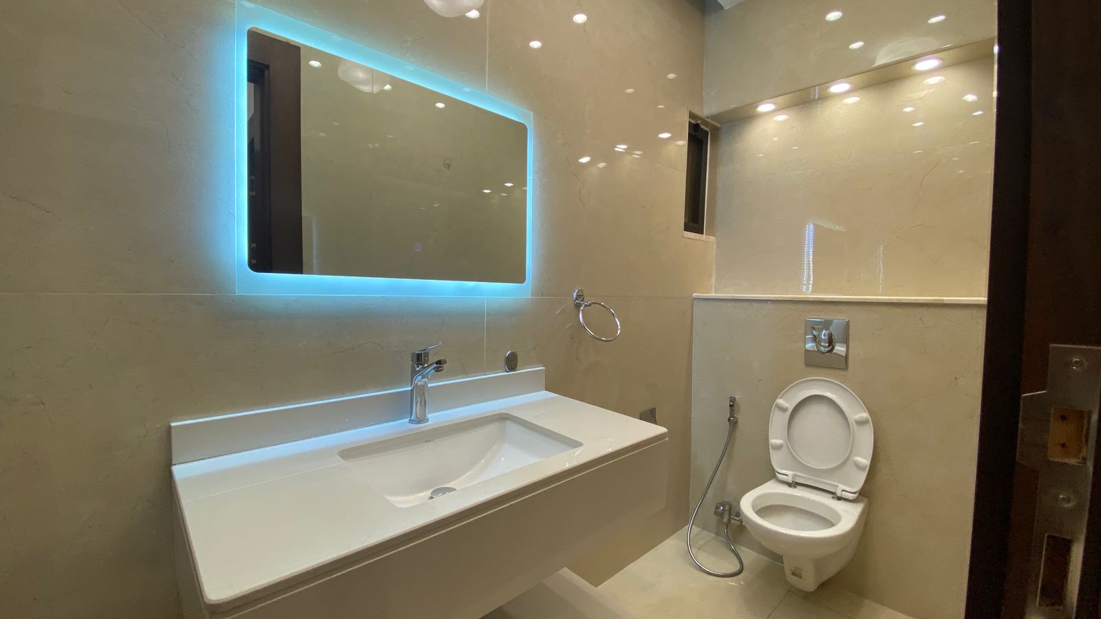 b7ef38a3-b0fb-4a3a-b19f-c2aa5a6bfea7 Modern bathroom with backlit rectangular mirror above a white sink, beige tiled walls, and a wall-mounted toilet with a bidet spray nearby.