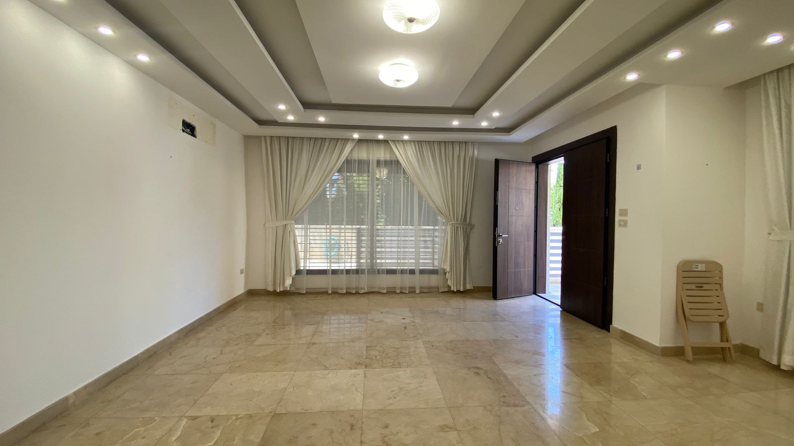 b884f148-4321-414e-b6e6-be2fd97a0020 Empty living room with polished marble tiles, white walls, and a large window with sheer white curtains.