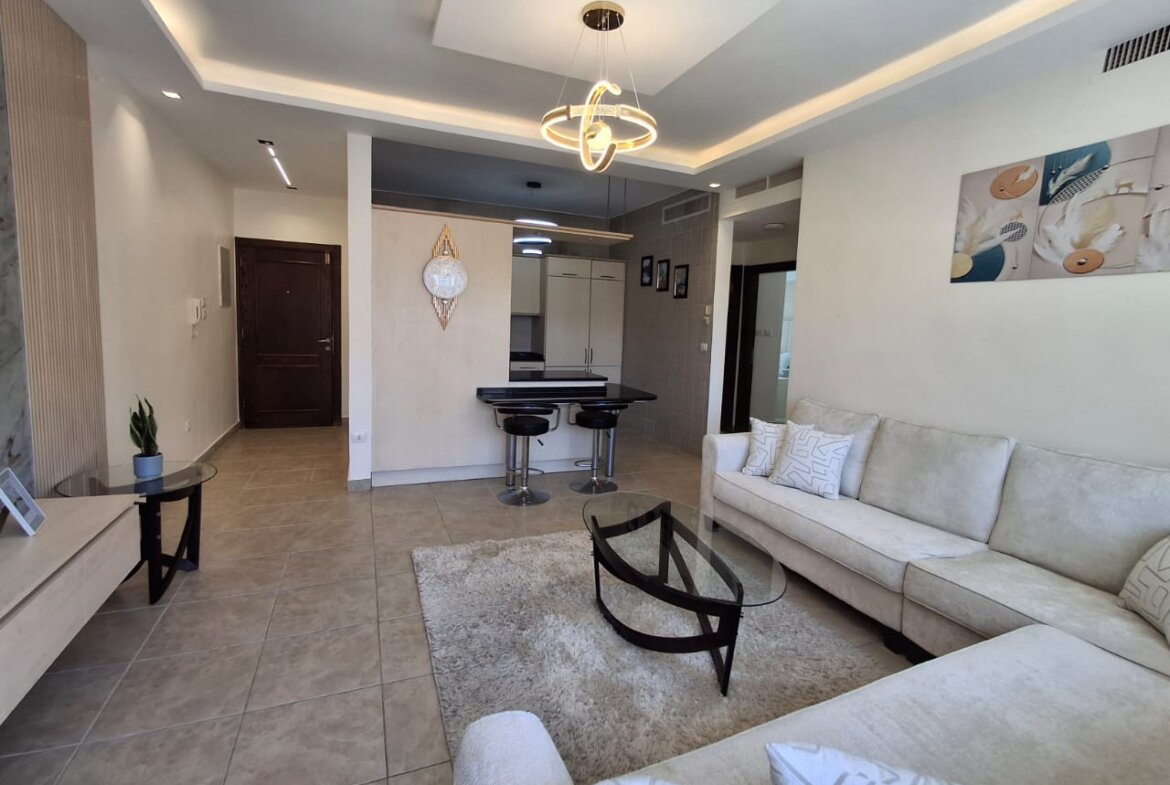 Open-concept living room with a cream sectional, glass coffee table, and shag rug facing a kitchen bar area with two stools and pendant lighting.