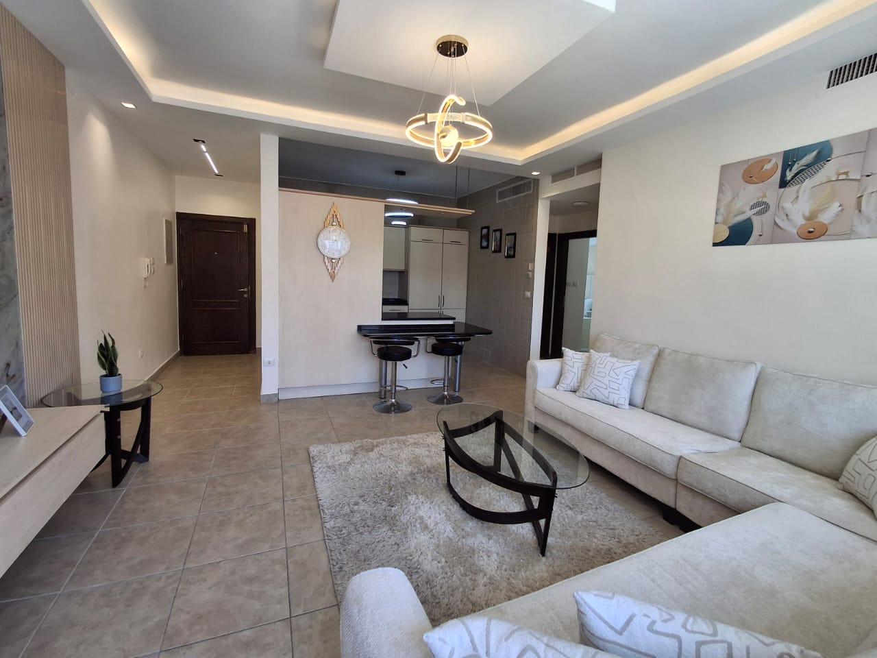 b8cdbdb7-0f68-41b3-8a91-3028710f49c2 1 Open-concept living room with a cream sectional, glass coffee table, and shag rug facing a kitchen bar area with two stools and pendant lighting.