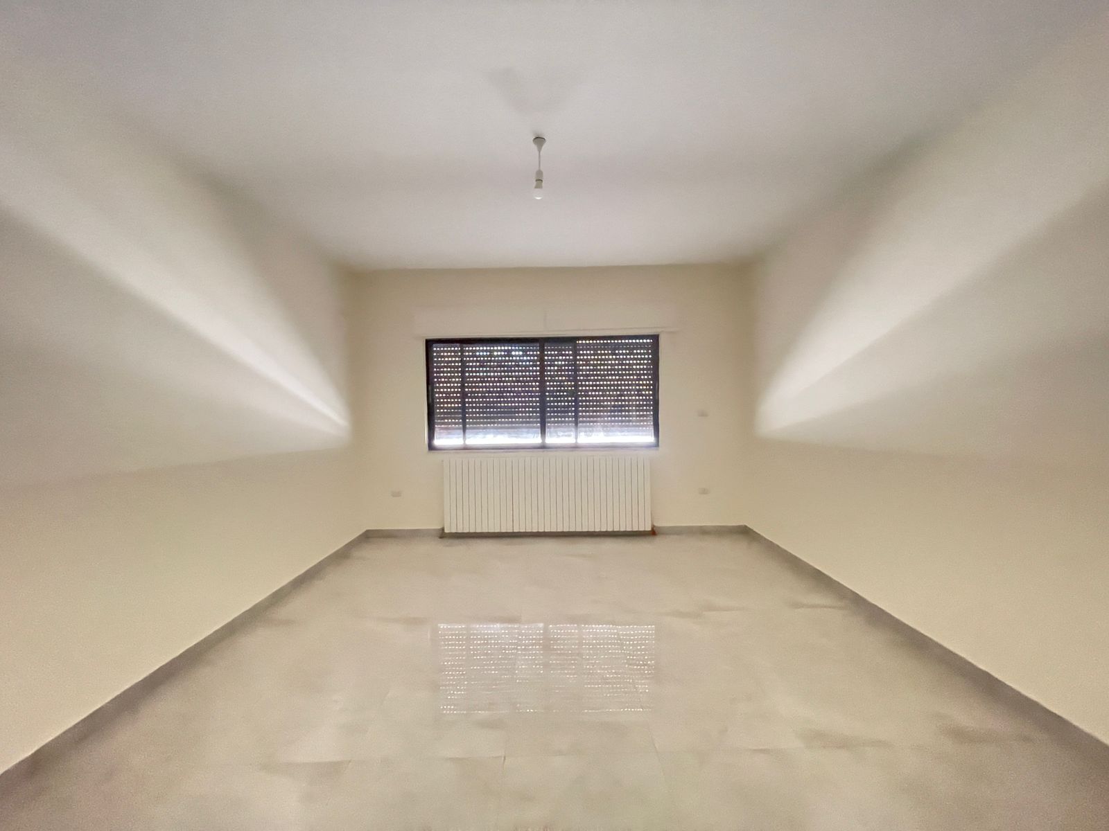 b92b281e-7edc-4511-ba31-ff2ded8dd3a2 Empty, unfurnished room with a single window and radiator beneath it; bare light bulb hangs from the ceiling.
