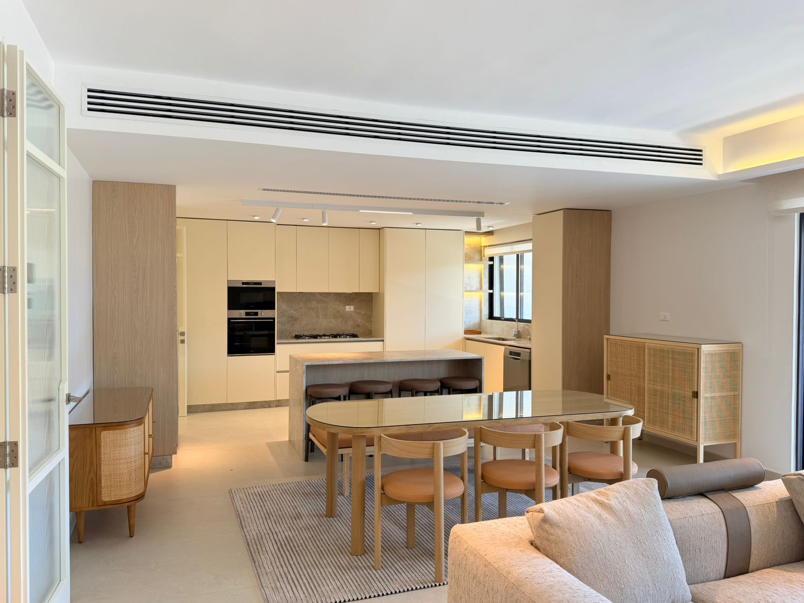 b92dc88f-36f1-465c-9dba-0152251f80bc Open-plan kitchen with beige cabinets, island and wooden dining table with six stools in a bright living area.