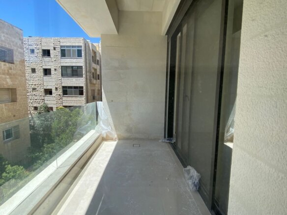 Balcony with glass railing overlooking neighboring beige stone buildings and a clear blue sky beyond.
