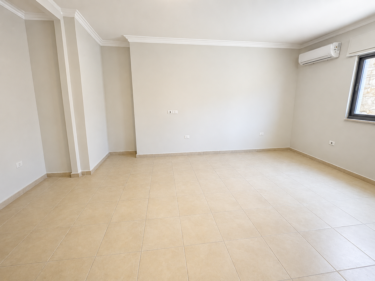 bac2a95e-e884-4d4b-81b9-9edb6479bc33 Empty beige-tiled living room with light gray walls, white crown molding, and a wall-mounted air conditioner; window on the right showing stone exterior.