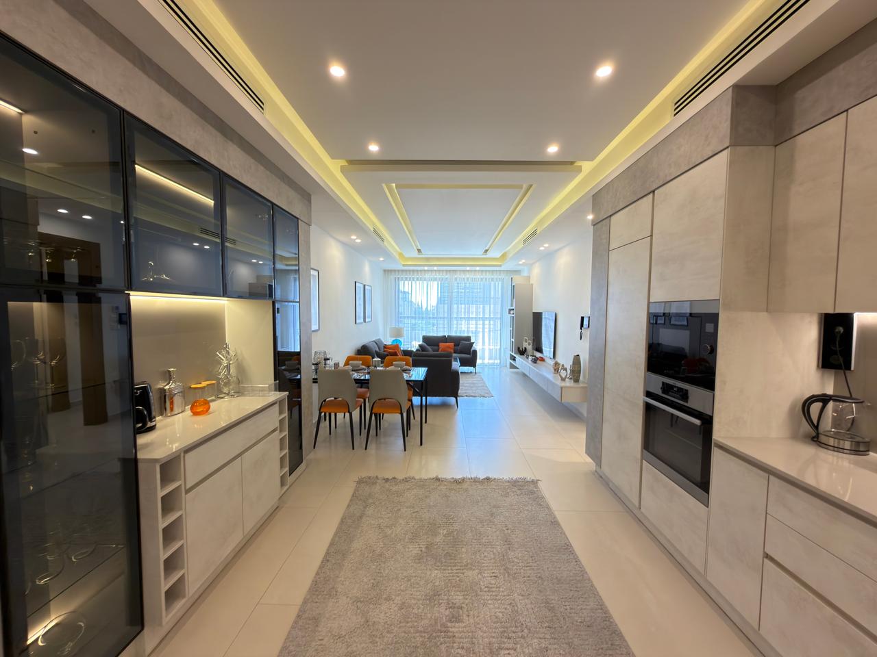 bac4c6d1-2380-4301-8557-79a7e16153d1 Open-plan kitchen and living area with gray cabinetry, built-in appliances, and a glass-fronted wall unit; dining table with orange chairs beyond a long gray rug.