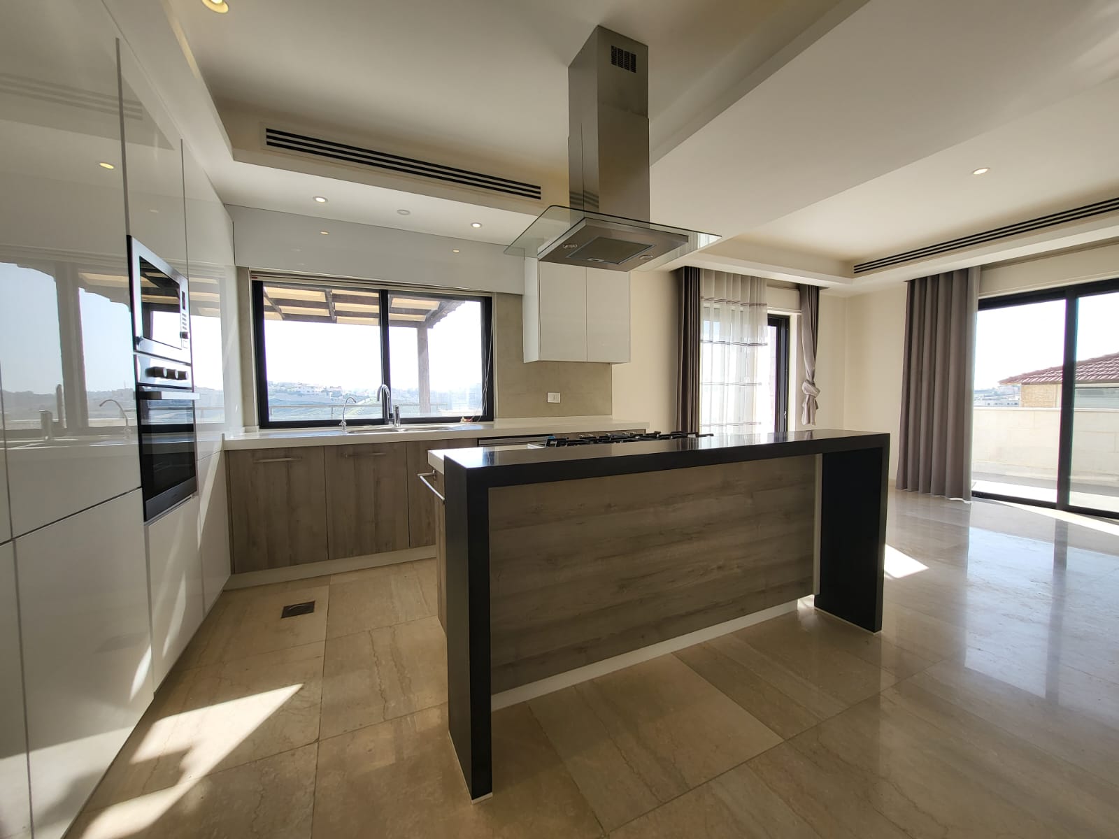 bcbbd395-f158-44ea-9564-7e24dbec142f Modern kitchen with an island, stainless steel range hood, and built-in oven; white upper cabinets and wood-toned lower cabinets under bright, sunlit windows.