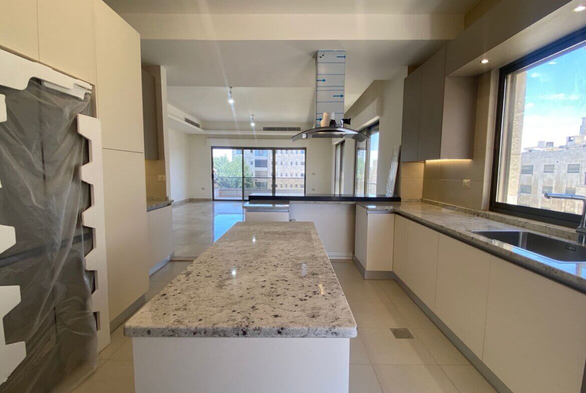 bd3dcc33-cb36-4d32-a77d-792d174afc35 Modern kitchen with white cabinets, granite countertops, and a central island; large windows overlook a cityscape. The tall appliance on the left is wrapped in protective plastic, indicating new setup.