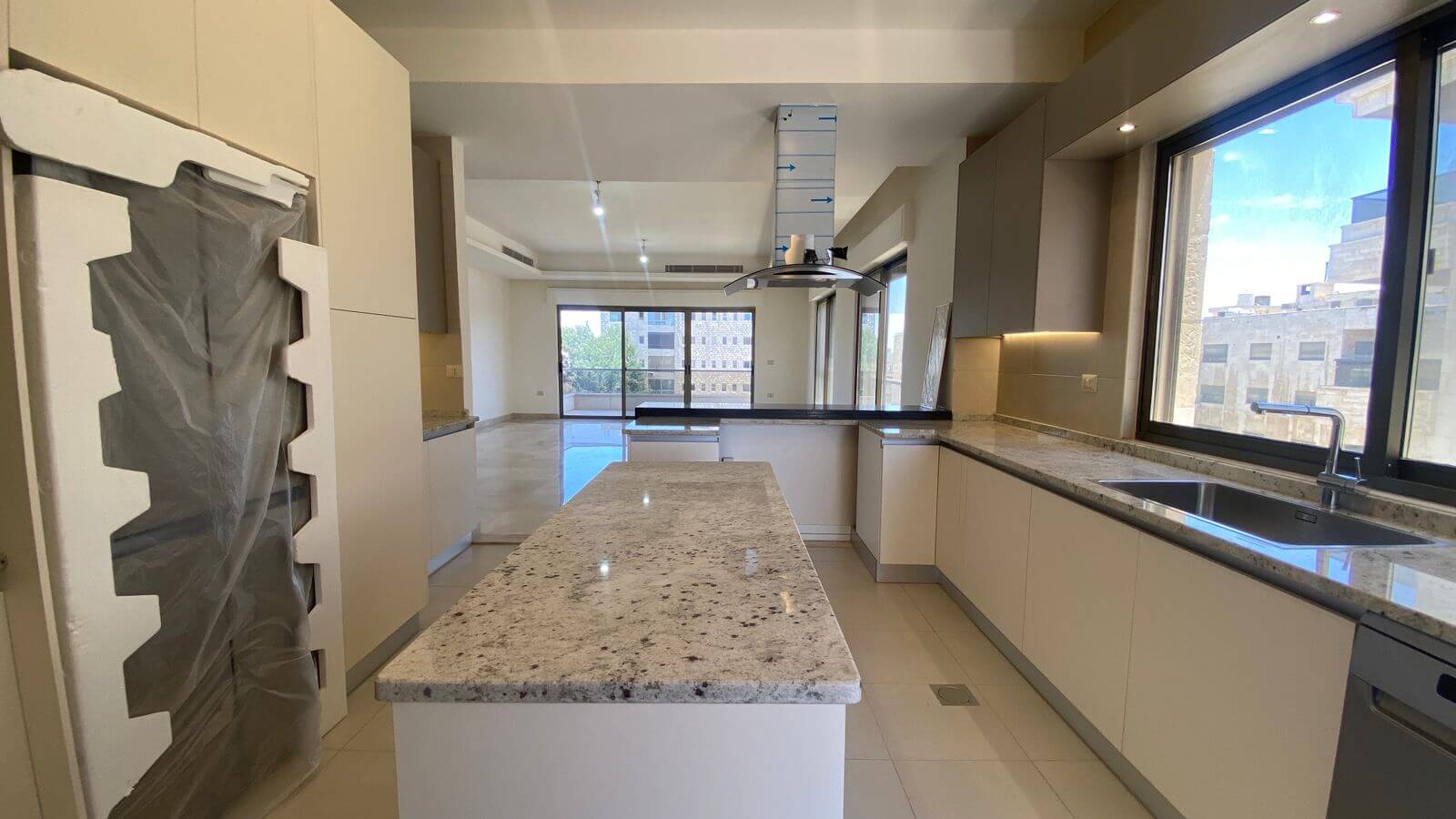 bd3dcc33-cb36-4d32-a77d-792d174afc35 Modern kitchen with white cabinets, granite countertops, and a central island; large windows overlook a cityscape. The tall appliance on the left is wrapped in protective plastic, indicating new setup.