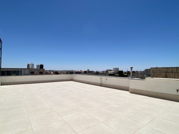 Wide rooftop terrace with beige tiled floor and a broom leaning on the wall, blue sky above and a distant city skyline.