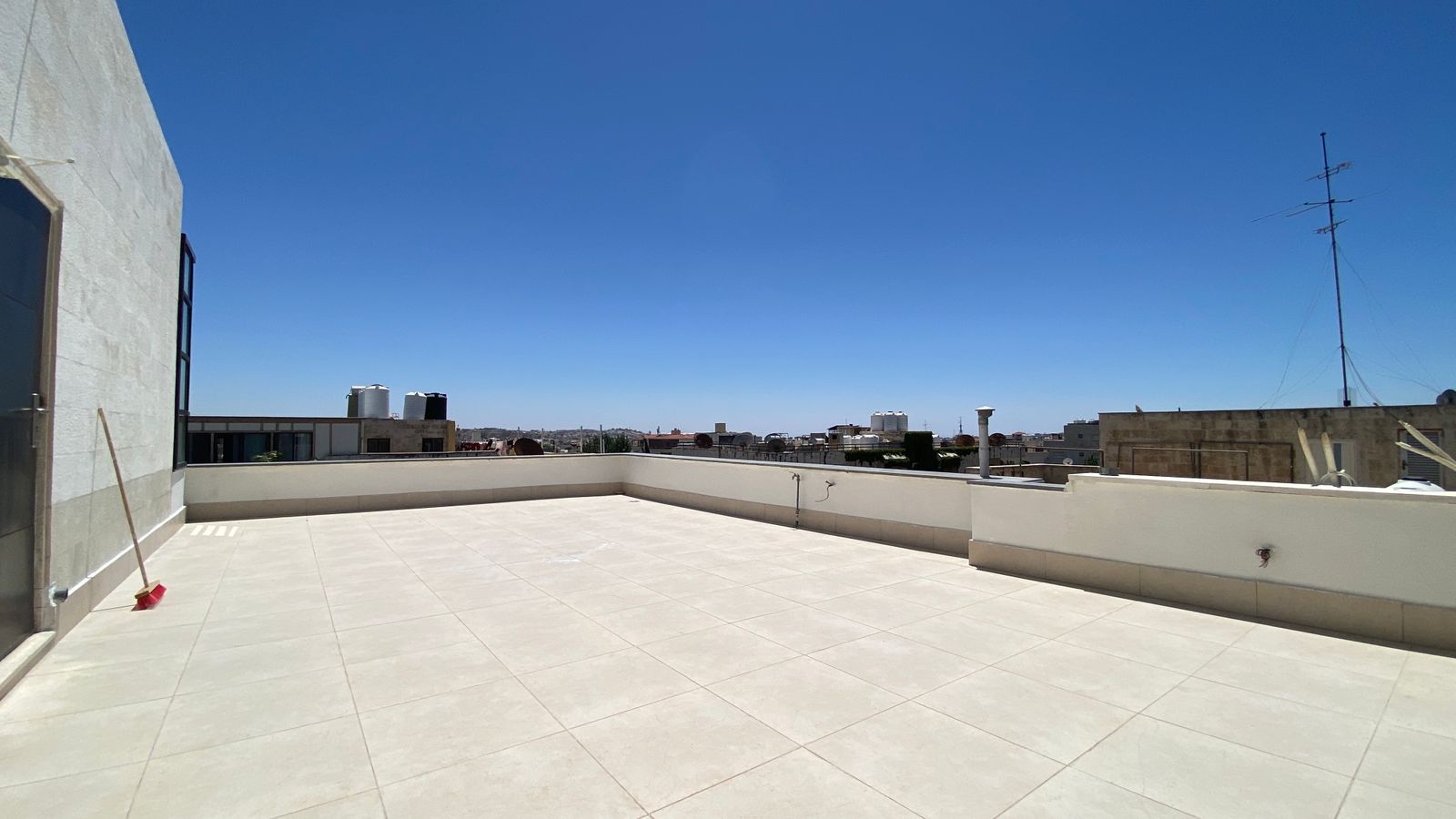 bde8ab17-05cd-499d-b8ea-a66d2c7813d9 Wide rooftop terrace with beige tiled floor and a broom leaning on the wall, blue sky above and a distant city skyline.