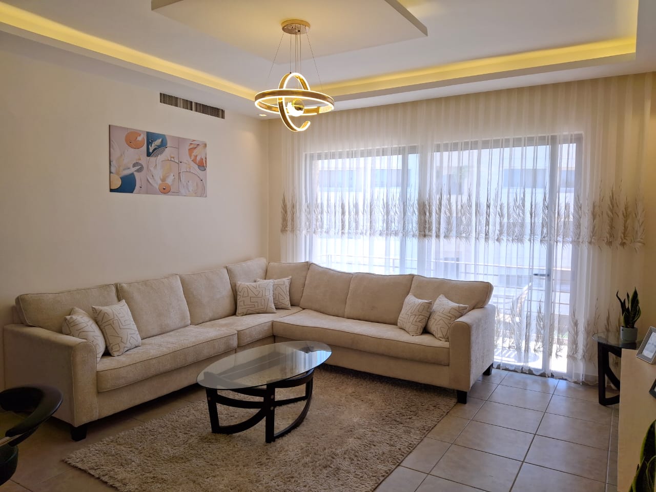 Living room with a beige L-shaped sofa, glass oval coffee table, and a cream rug bathed in natural light from large sheer-curtained windows.