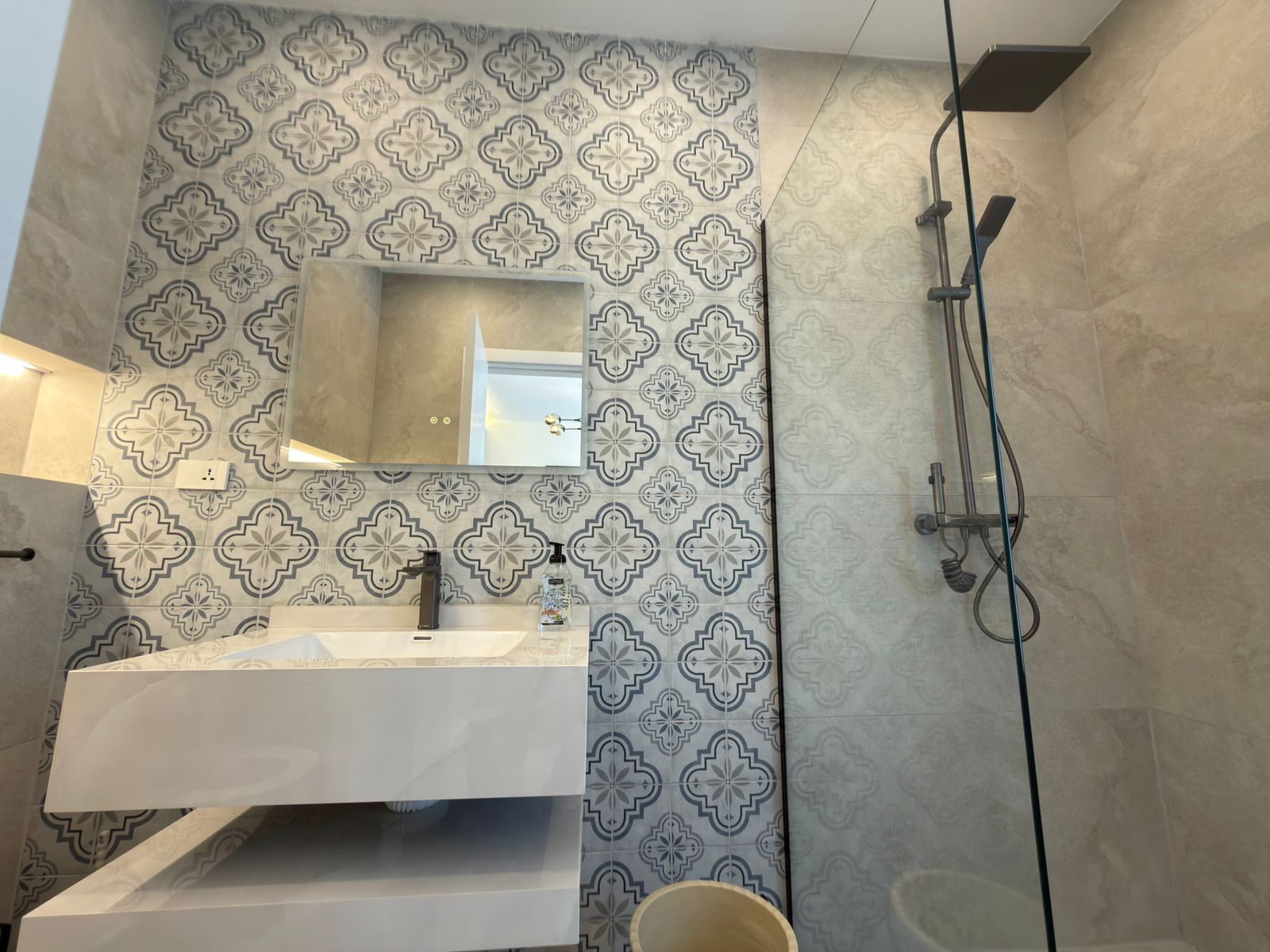 bedc77dc-879b-47a9-baa8-af0024575b17 Modern bathroom with a blue-and-white patterned tile wall, a white vanity with a black faucet, and a glass-enclosed shower with a rainhead.