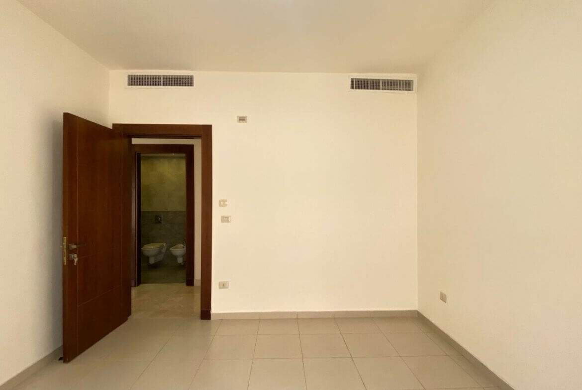 beef8d30-b8e7-4b42-8d3f-55d2151b3dc1 Empty, neutral-toned room with tiled floor; a wooden door is ajar, revealing a small bathroom with two toilets beyond.