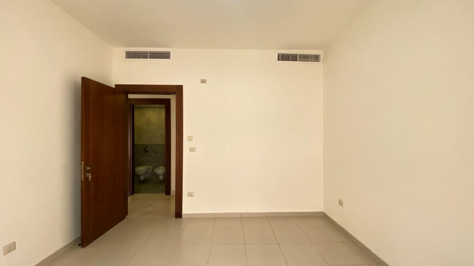 beef8d30-b8e7-4b42-8d3f-55d2151b3dc1 Empty, neutral-toned room with tiled floor; a wooden door is ajar, revealing a small bathroom with two toilets beyond.