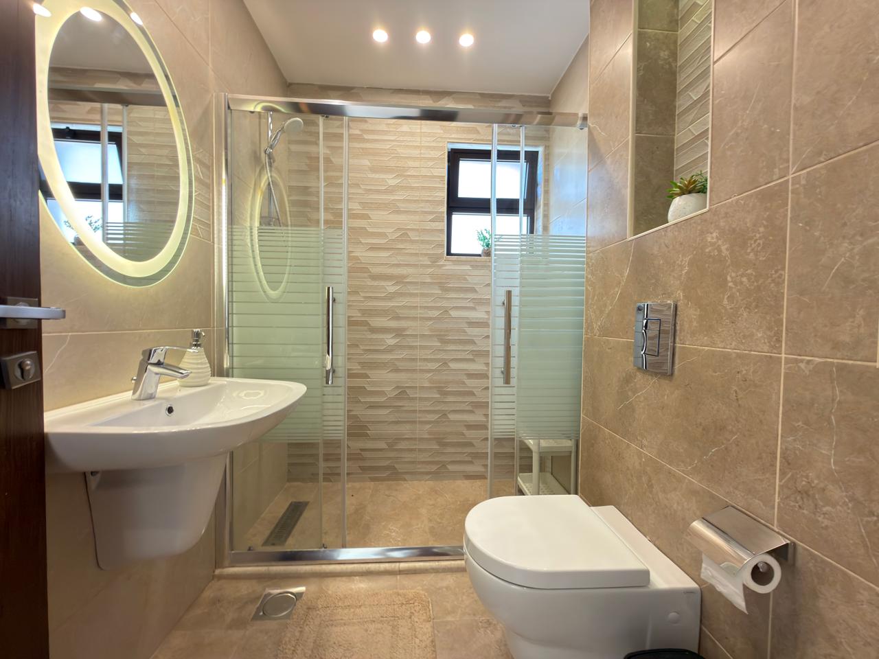 bf0ad489-de03-4516-b5d6-e91b274771c9 Modern beige bathroom with a wall-mounted sink, backlit oval mirror, glass shower enclosure, and a wall-hung toilet with a chrome paper holder on the right.