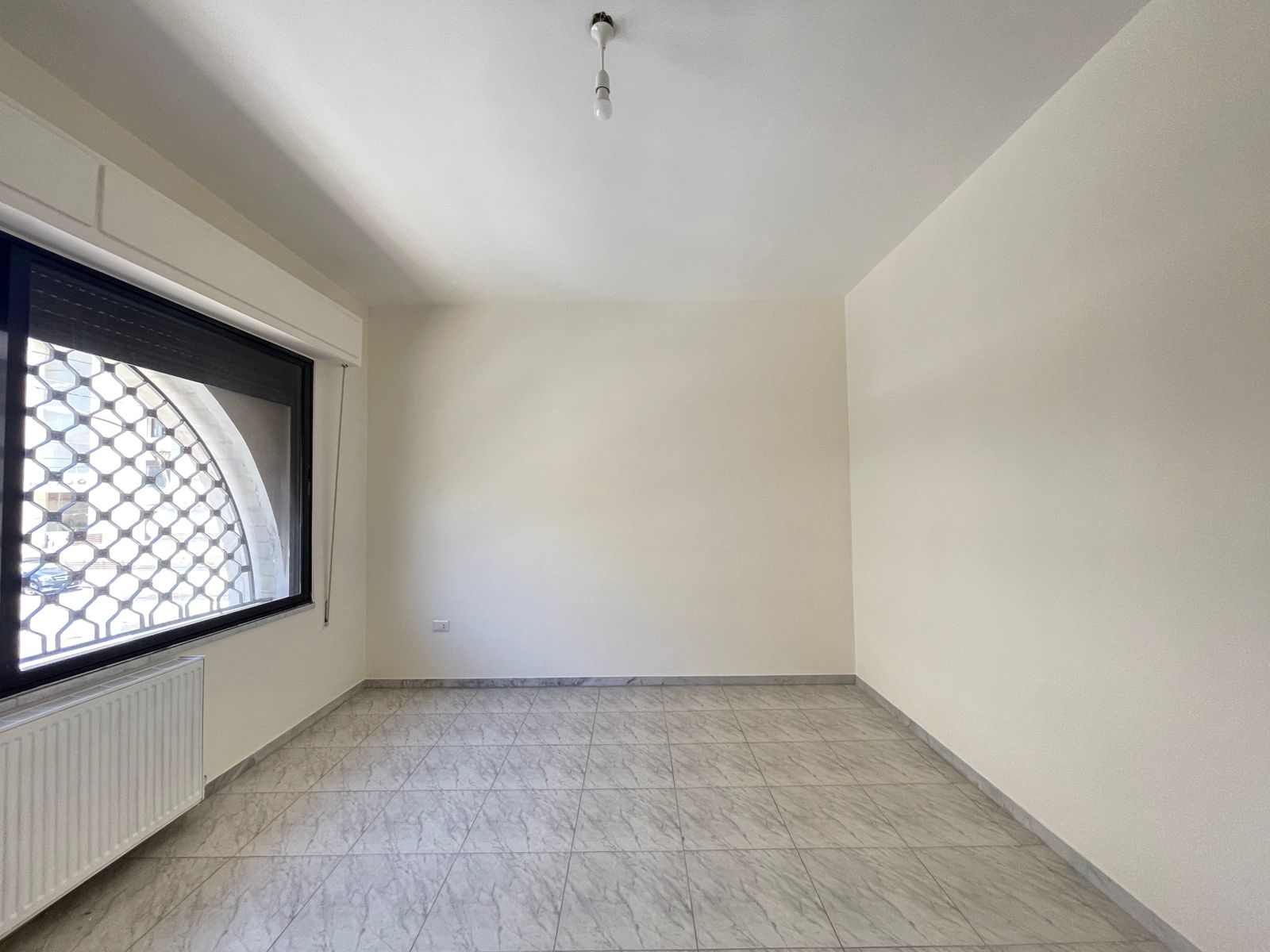 bf0fffa7-8c52-40e9-aff9-02930e501e3b Empty room with white walls and tiled floor, large window with decorative metal grille, radiator below, and a bare hanging light bulb.",