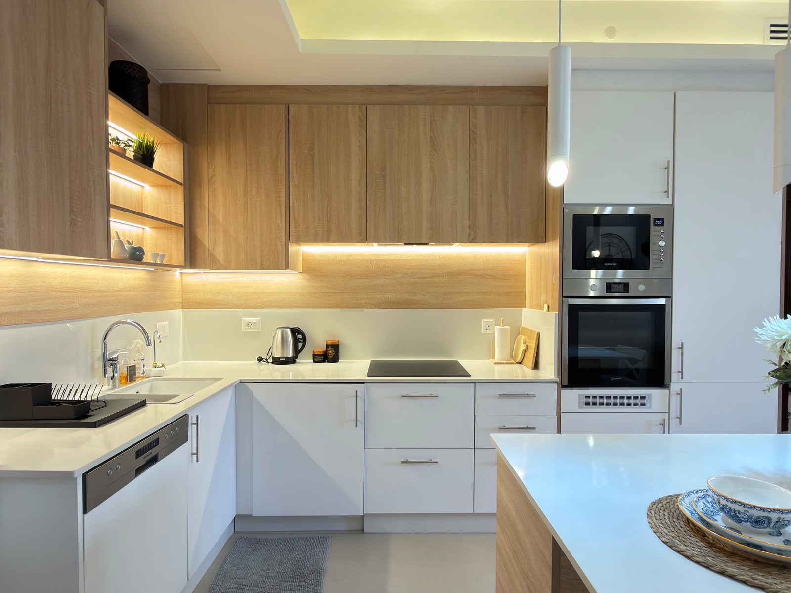 c0bf8612-38a6-490b-93b0-b530d891eae3 Modern kitchen with white lower cabinets, wood upper cabinets, under-cabinet lighting, and a built-in oven/microwave stack on the right.