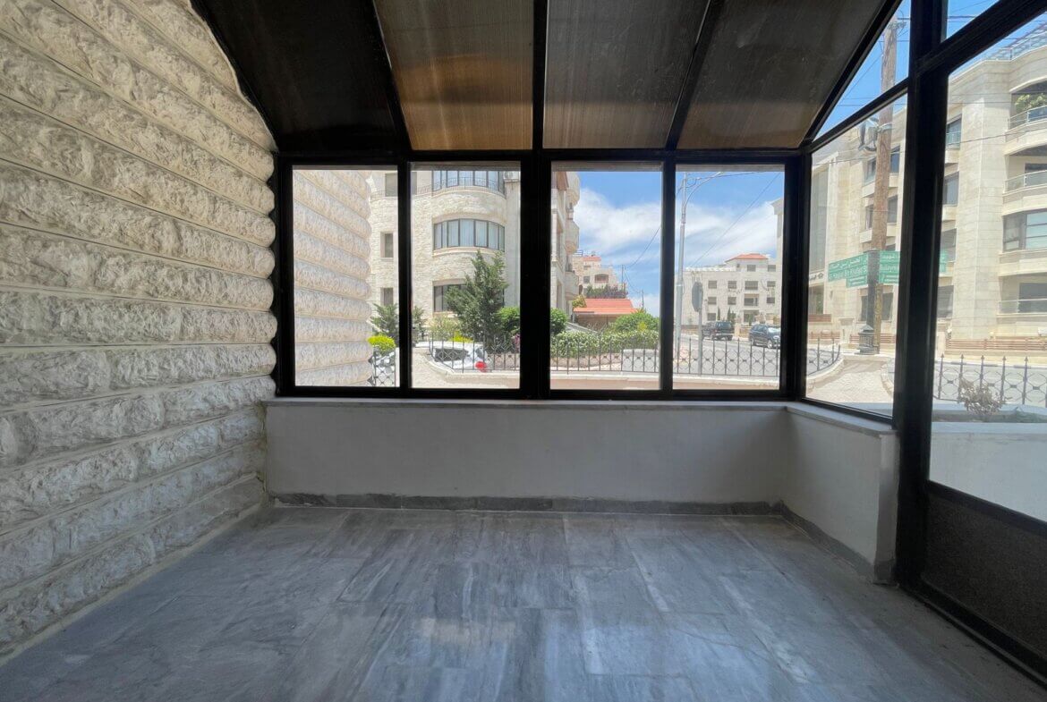 c179d117-b1b7-493c-bf80-3864d12cc759 Enclosed balcony with textured stone walls and large windows looking out on a city street with trees and modern buildings.