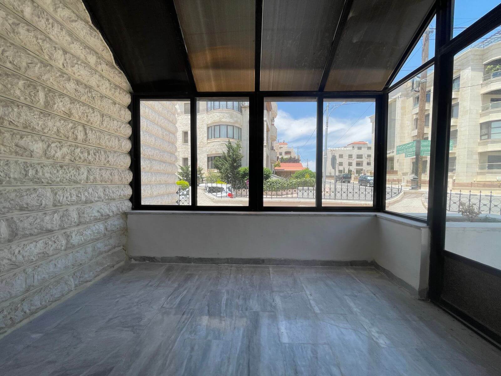 c179d117-b1b7-493c-bf80-3864d12cc759 Enclosed balcony with textured stone walls and large windows looking out on a city street with trees and modern buildings.