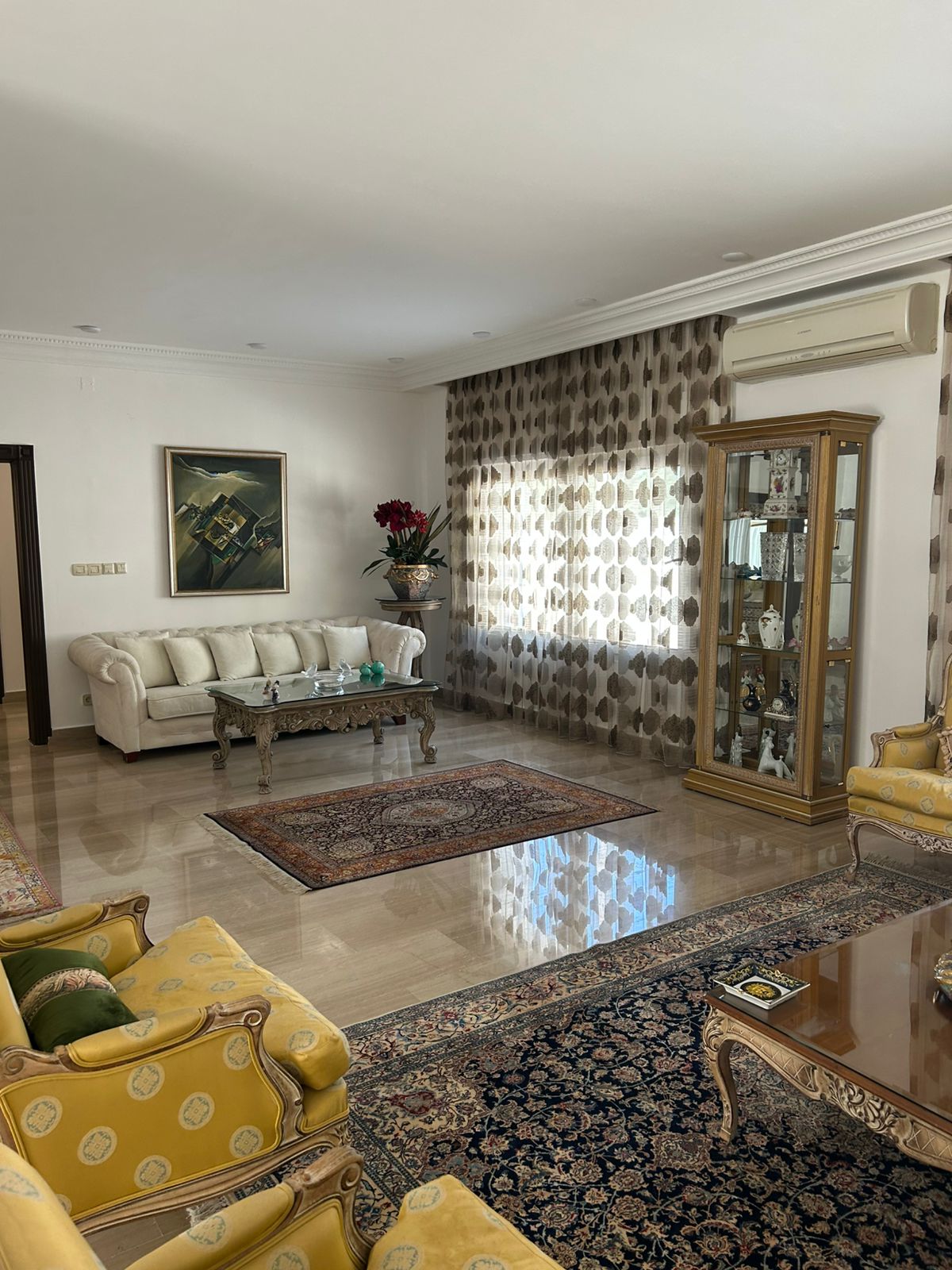 c20166d9-7dea-4b63-b845-483040a07f3d Elegant living room with a cream sofa, ornate glass coffee table, and patterned curtains beside a glass display cabinet.
