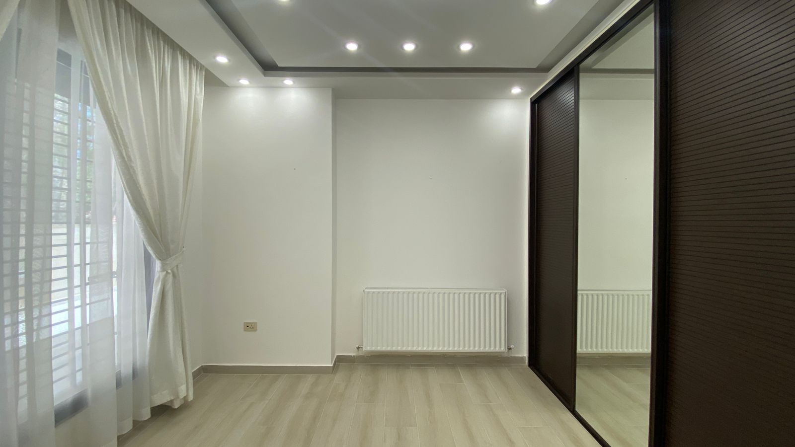 c3386ee6-75df-4cec-9be1-2b9028ae4c77 Bright, empty room with white walls, sheer curtains by a window, a central radiator, and dark sliding mirrored closet doors on the right.