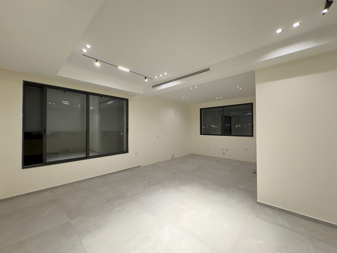 c3ac336b-ba15-47d0-9dae-e8dfc59f0491 Empty modern living space with white walls, gray tile flooring, and large black-framed windows. Recessed ceiling lights illuminate the room at night.