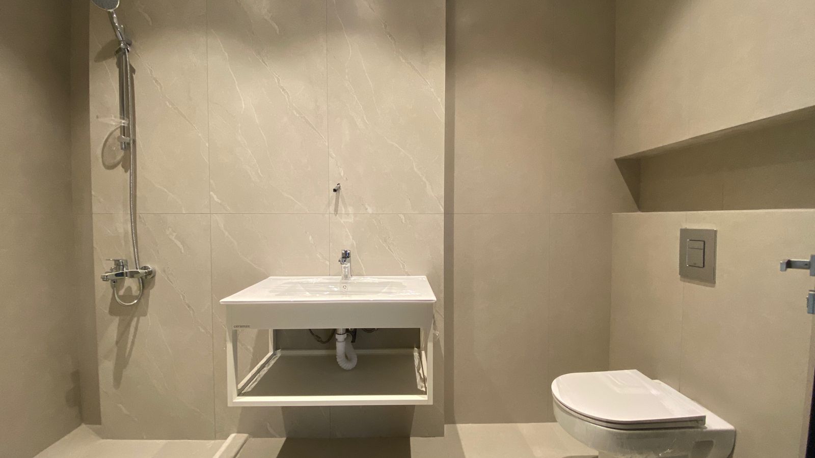 c48a7d29-2ecd-4434-aae9-9e9999e32b65 Modern beige-tiled bathroom with a wall-mounted sink centered under a mirror area, a chrome showerhead and handheld shower on the left, and a wall-hung toilet on the right.