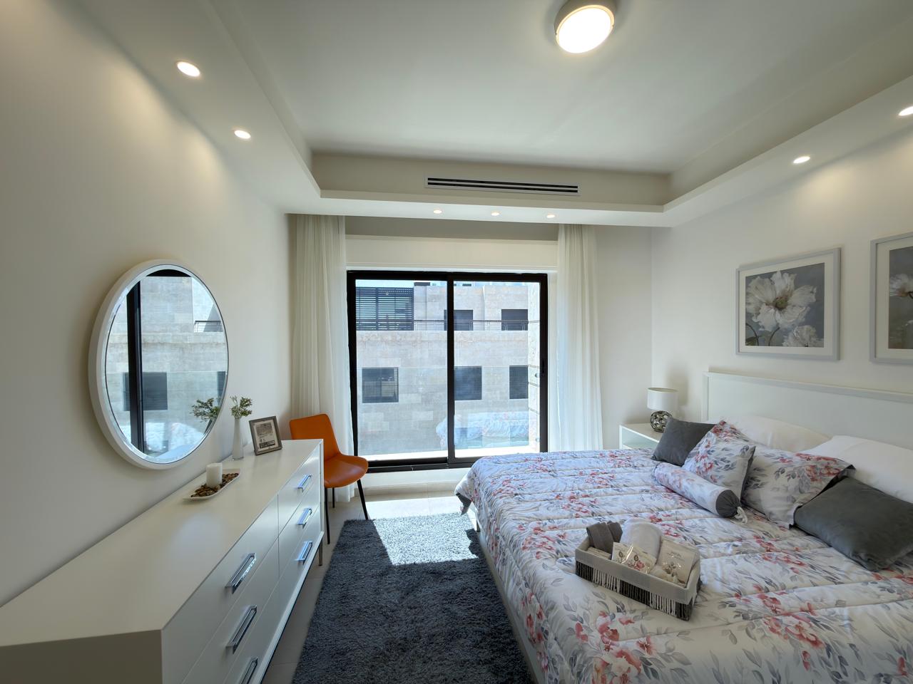 c4bf8d62-04fd-4c6d-9e34-b36bcea7578c Bright modern bedroom with white dresser, round mirror, and bed covered in floral quilt facing a large sliding glass door with a city view.