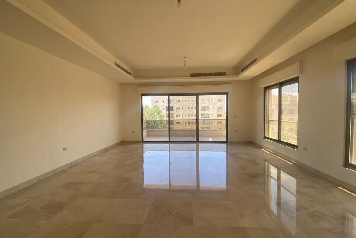 c4c6c887-28b1-4571-baf7-8036b84cac43 Bright empty living room with marble tile floor, neutral walls, and large sliding glass doors to a balcony showing nearby buildings.