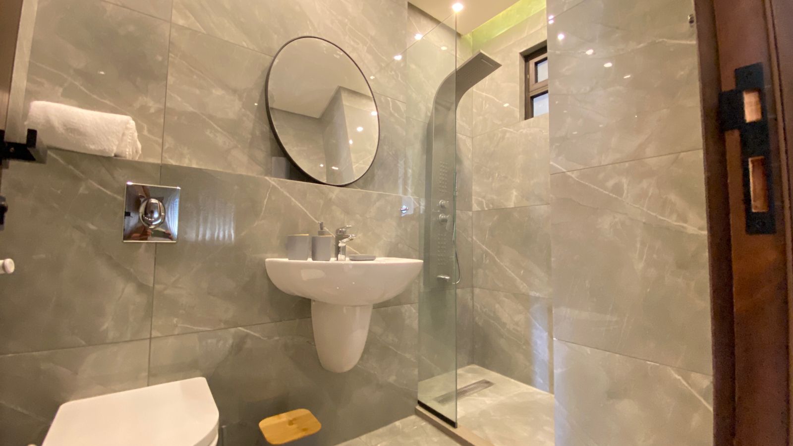 c4f40b37-7330-43ee-80af-319dde08b6cf Grey marble bathroom with wall-mounted sink, round mirror, toilet, and glass-enclosed shower - modern and minimalistic interior.