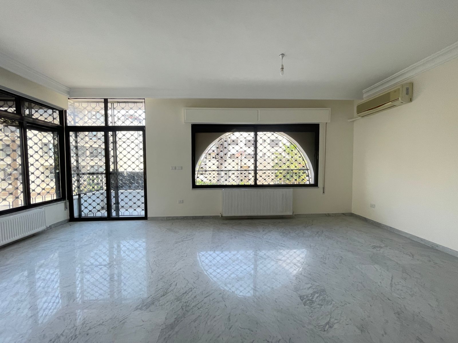 c5d359f2-7b01-4956-a752-70317958f5a4 Bright empty living room with marble floor, large arched window with metal grill, radiator below, and a glass door to the left; wall AC unit on the right.