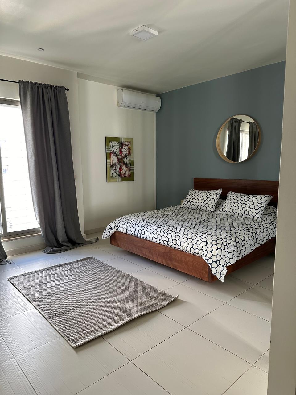 c5dd0fee-85e7-48f5-b8b0-0633b40721c3 Modern bedroom with a wood platform bed, geometric bedding, blue accent wall, and a round wall mirror above the headboard.