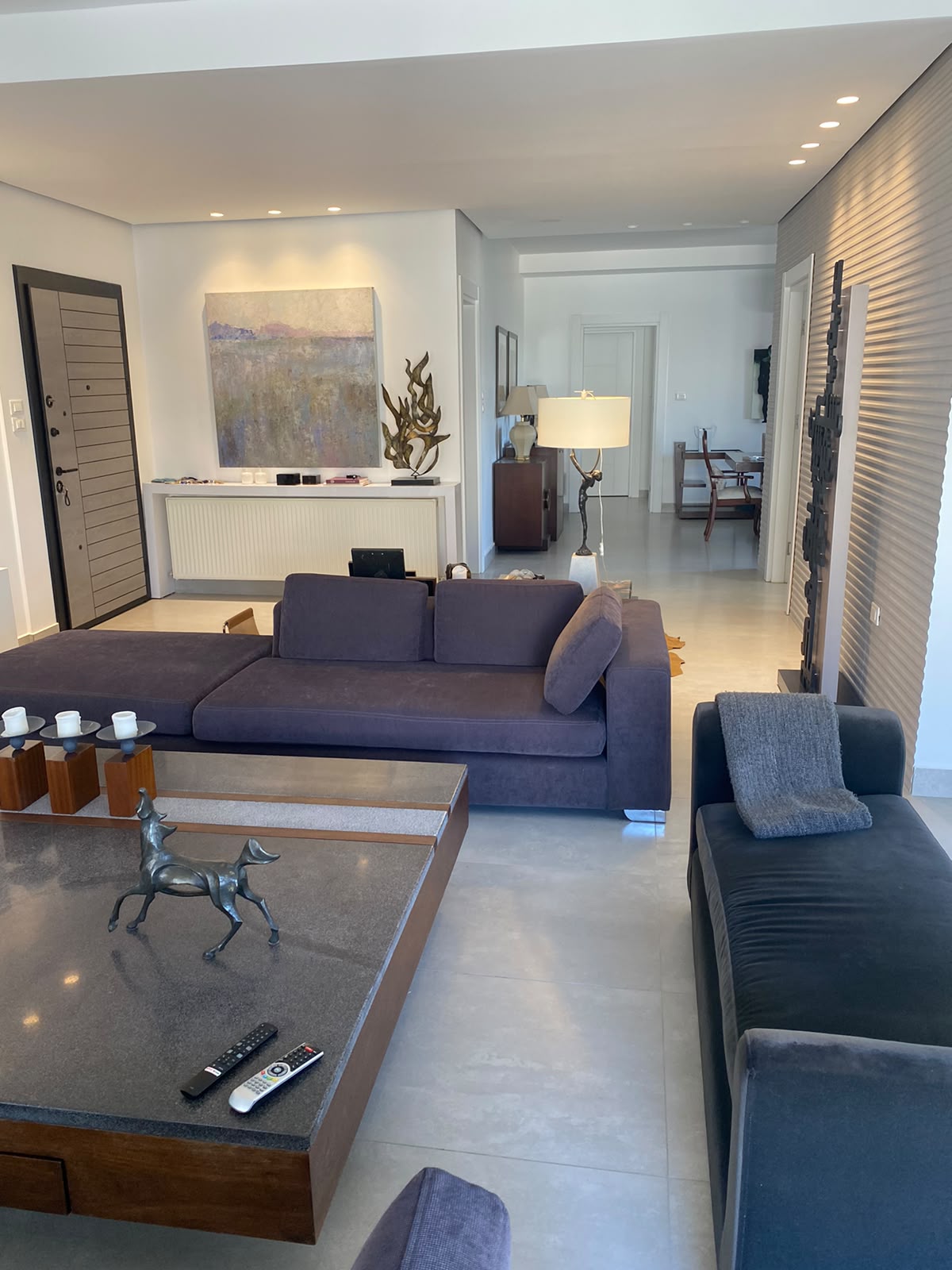 c5f59b33-3c50-4985-9d4b-fe5afe22ac10 Modern living room with a dark purple sectional sofa, glass-topped coffee table, decorative horse sculpture, and abstract wall art above a radiator.