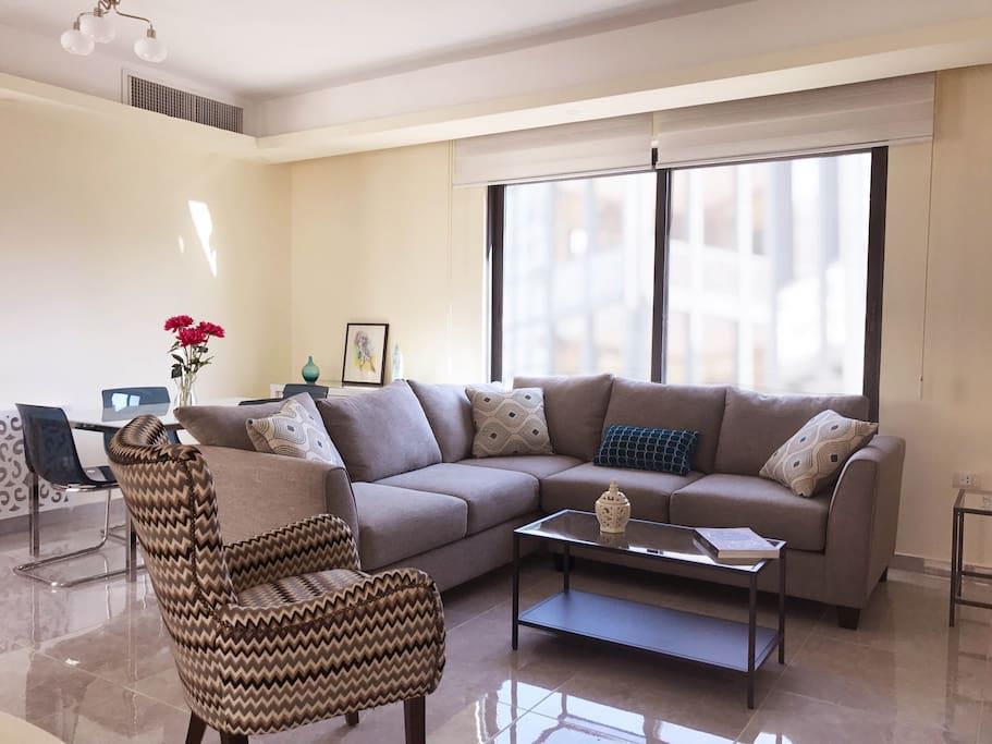 Modern living room with a light gray sectional sofa, a patterned armchair, and a glass coffee table by a large window.