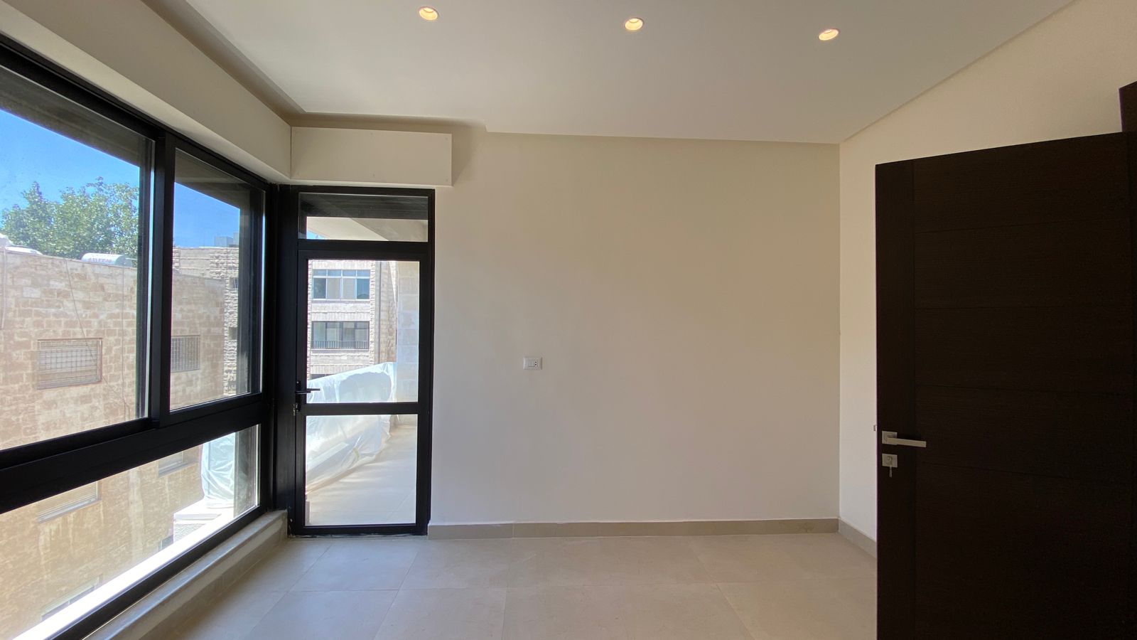 c77db20c-1815-4bec-90ab-b323714739c0 Empty room with large black-framed window and glass balcony door, beige tile floor, white walls, and a dark wood door on the right.