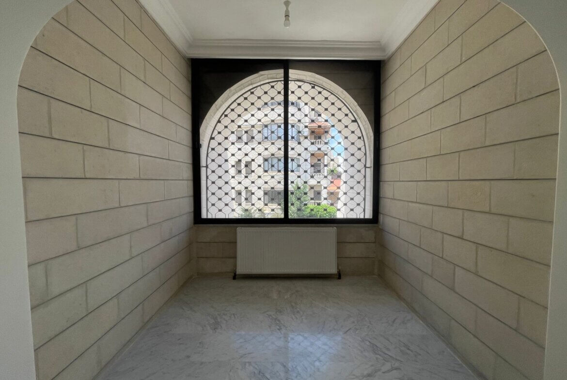 c80cb07d-34fe-47e8-b6c5-d3baa0fc6231 Narrow indoor hallway with light beige stone walls, an arched window with a metal lattice grille, and a radiator beneath it.