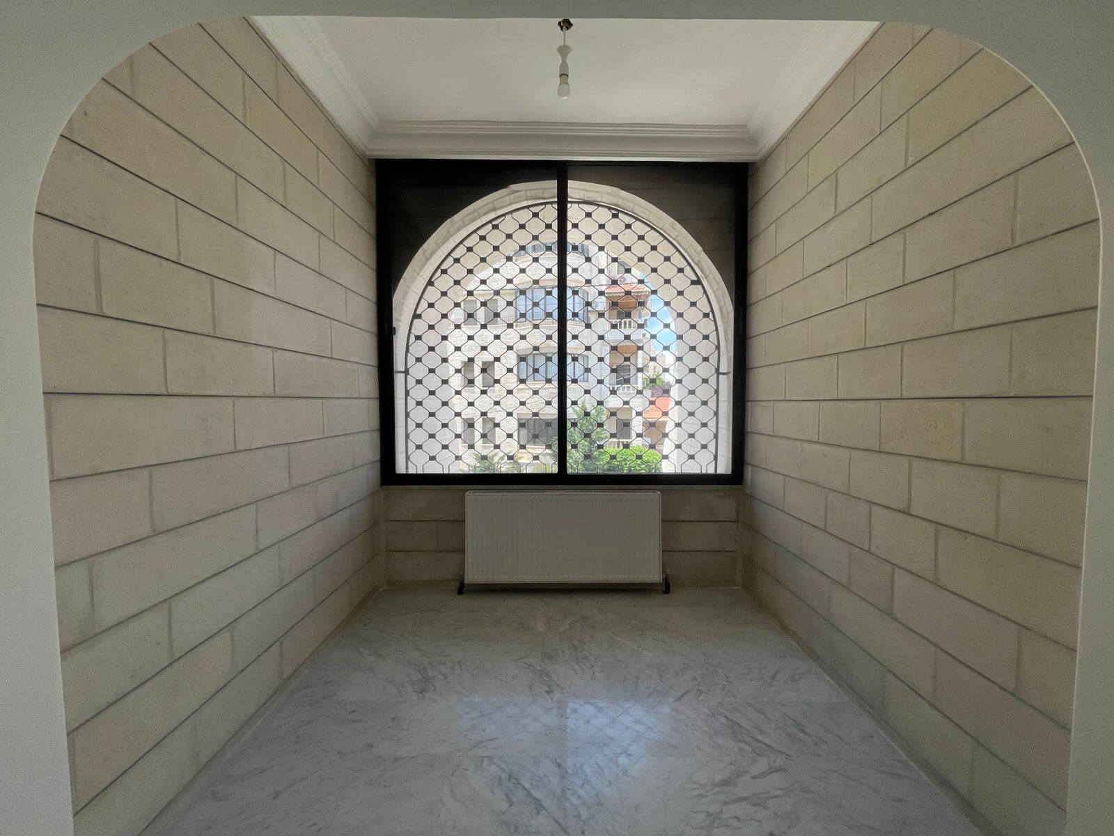 c80cb07d-34fe-47e8-b6c5-d3baa0fc6231 Narrow indoor hallway with light beige stone walls, an arched window with a metal lattice grille, and a radiator beneath it.