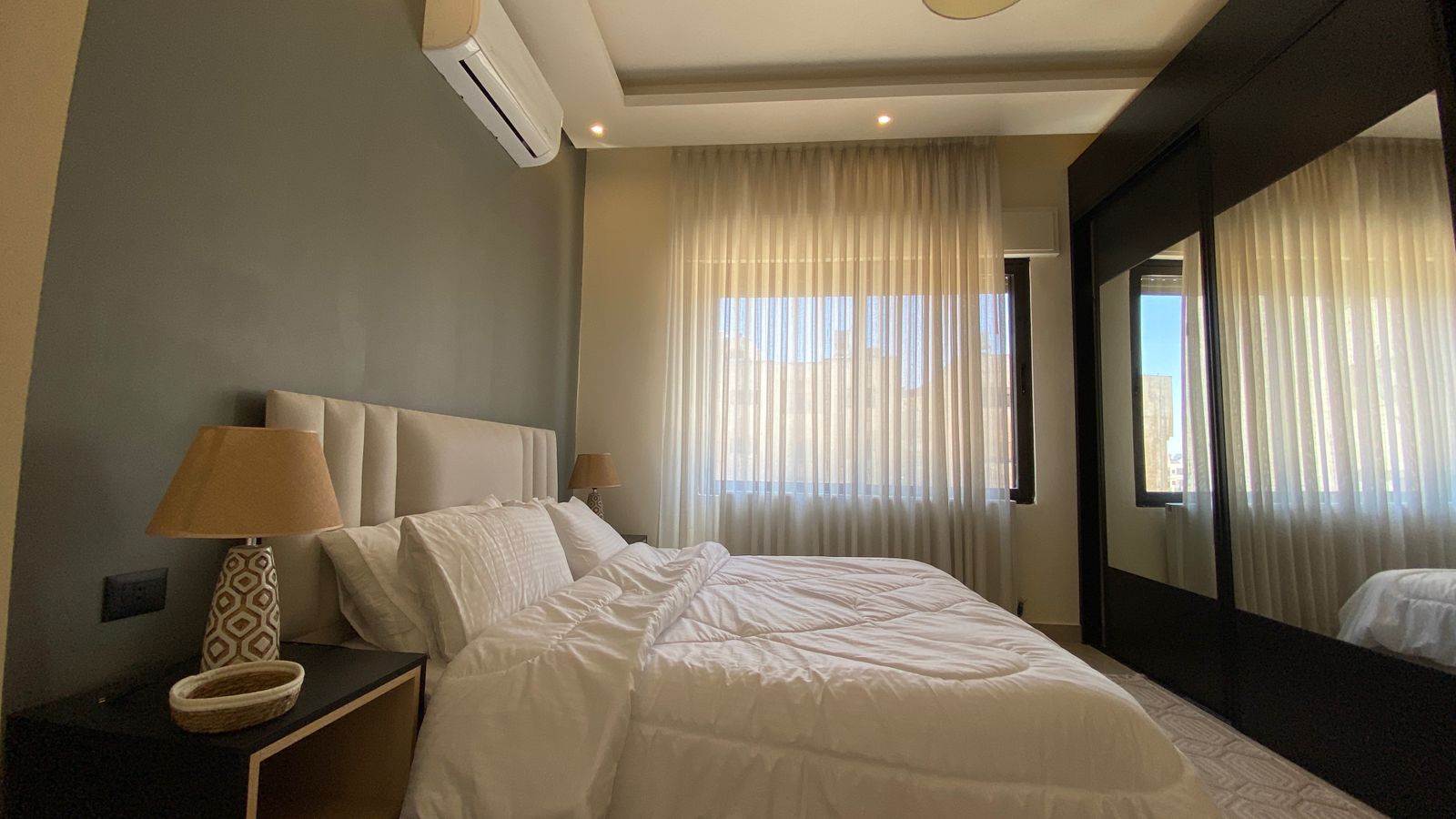 c8c7d920-0b6b-4267-b5ee-b10859598a79 Modern hotel-style bedroom with a large white bed, tufted headboard, and two beige lamps on dark nightstands by the window.