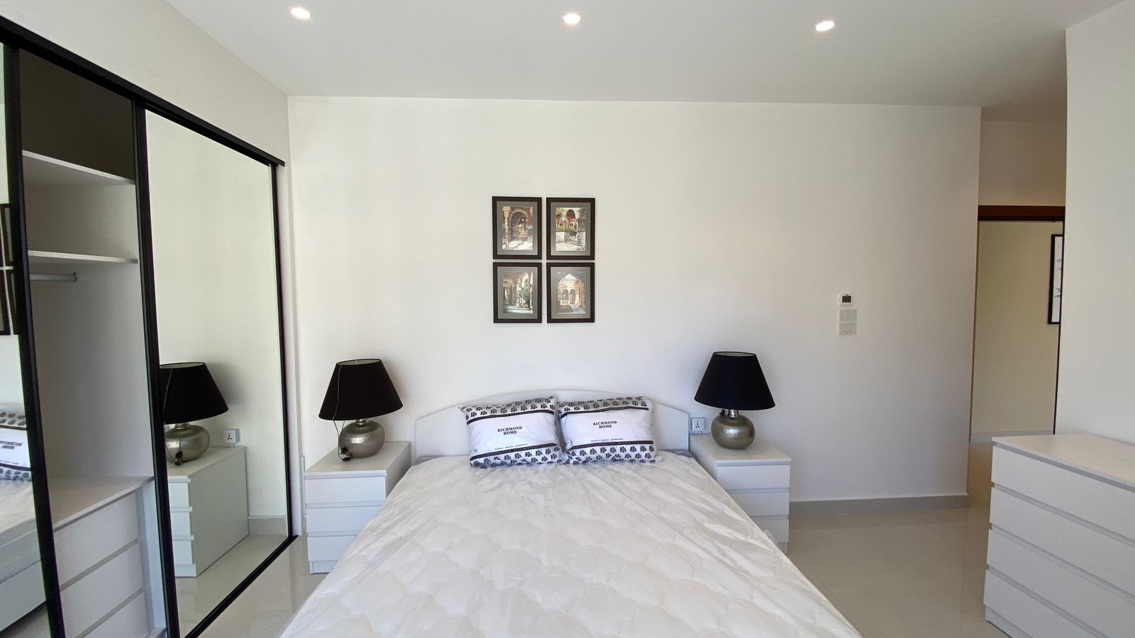 ca1f6848-1af1-48e8-91ae-b695abc77221 Minimalist white bedroom with a double bed, two nightstands, black lamps, and four framed pictures above the headboard.