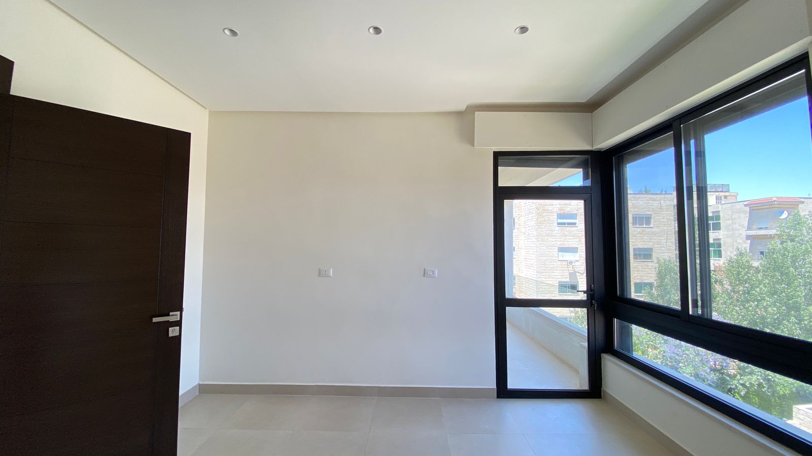 cac6ba4d-7e37-410b-9afe-8ab24bc48f07 Empty modern room with darkwood door, large windows, and balcony door letting in bright daylight.