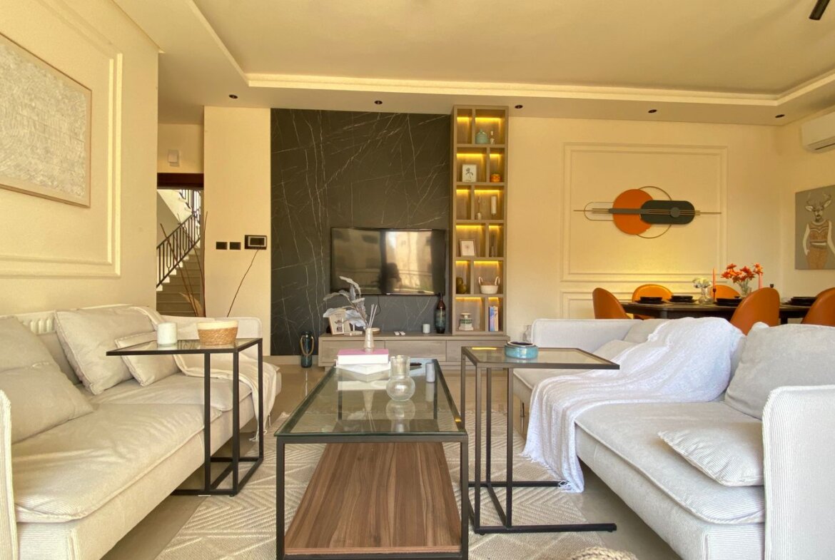 Modern living room with beige sofas, glass coffee tables, and a TV mounted on a dark marbled wall.