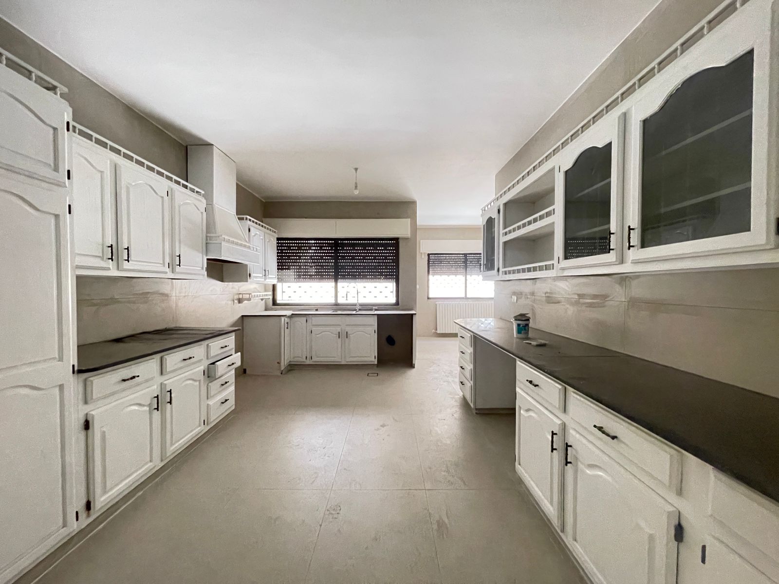 cb9f97b0-b2b7-435a-9186-bd70a6b953a7 Empty white kitchen with extensive cabinetry, dark countertops, and large windows with blinds at the far end.