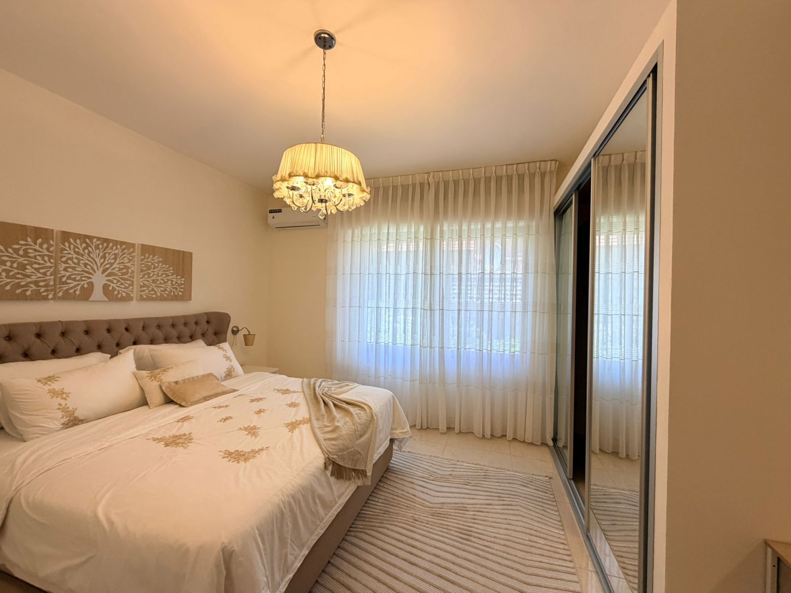 cbe4337b-acf6-4693-a4a7-03d6d5f9ddee Bedroom with a tufted headboard, white bedding with gold embroidery, and a hanging chandelier above the bed.