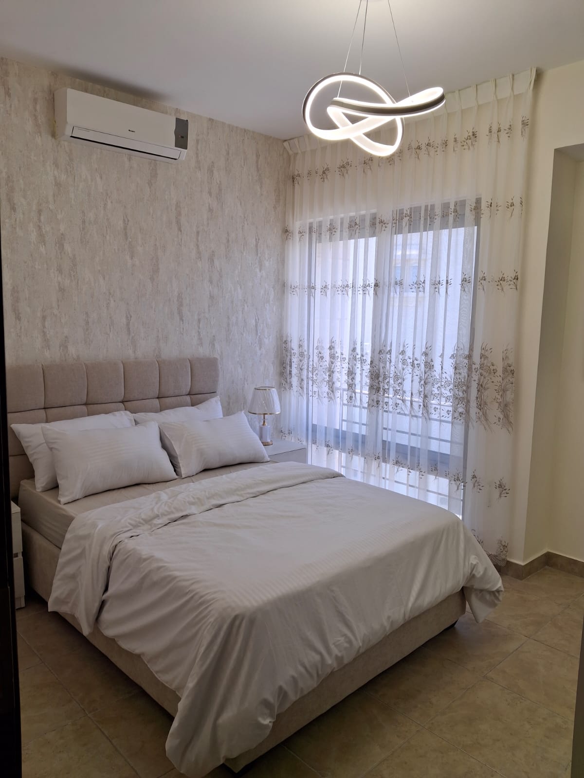 cc7a9c11-6955-4d4d-be38-5469ec5a9a48 Neutral bedroom with a beige upholstered bed, white linens, sheer floral curtains, and a sculptural loop ceiling light.