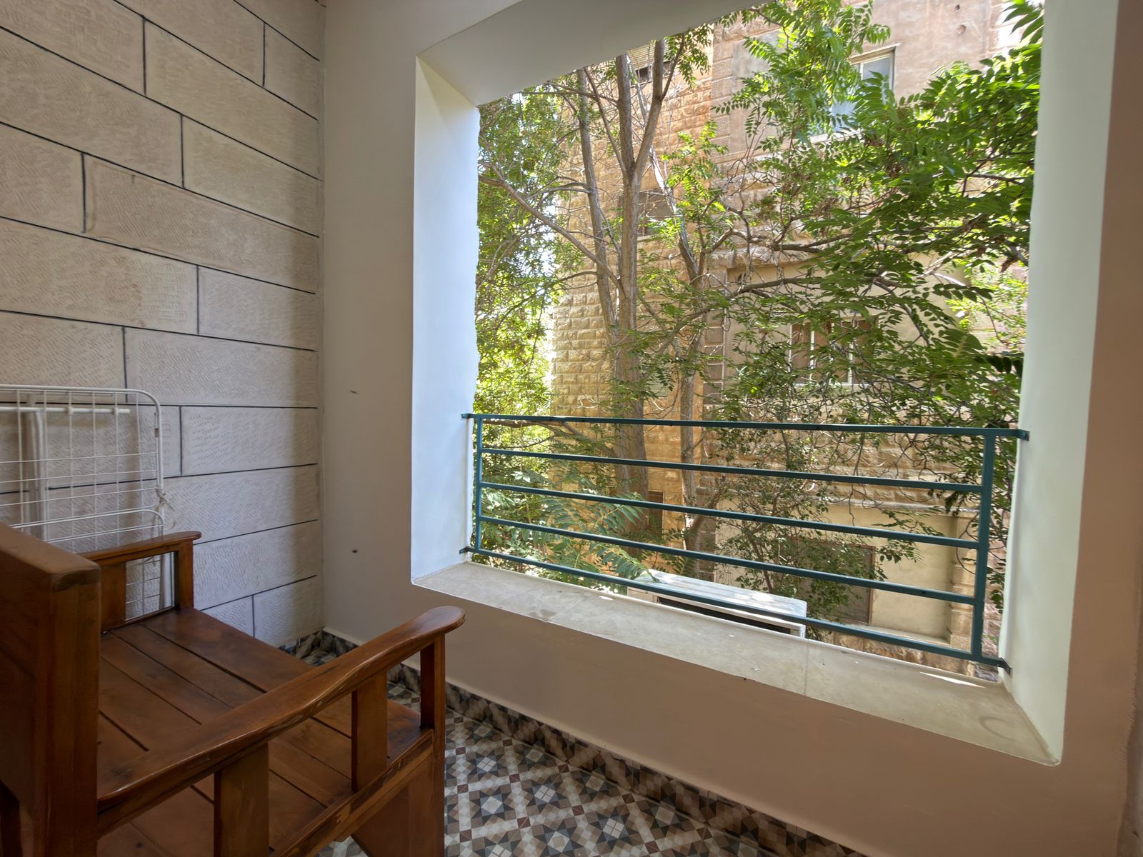 cd0ff13f-55de-4c2c-ab4b-b3d6444e72c4 A wooden bench on a narrow balcony with geometric tiled floor, stone wall left, and green trees beyond a metal railing outside the opening