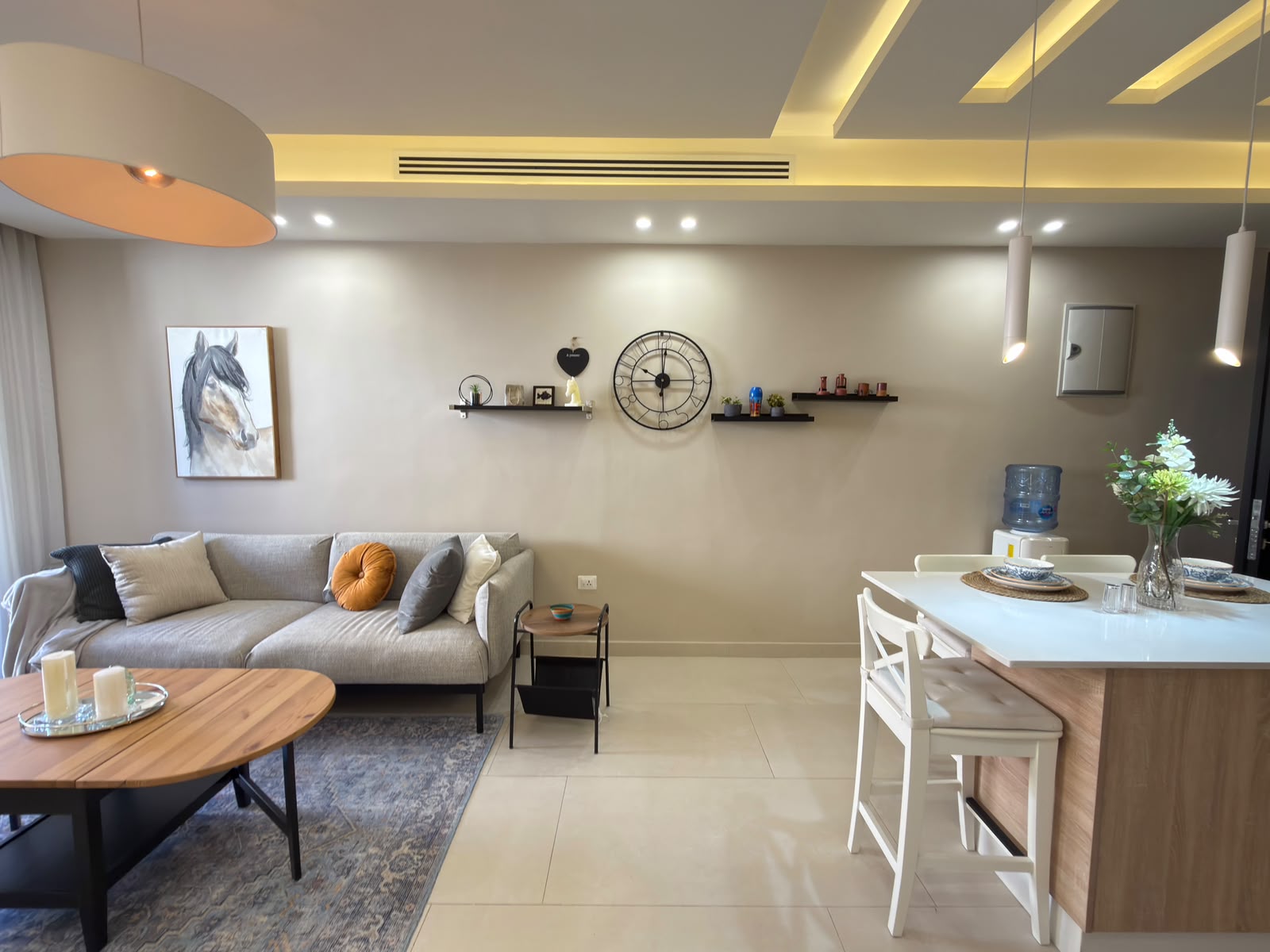 ce6a65b5-3687-49fd-9687-afe0dccfad1c Living room with light gray sofa, throw pillows, horse painting on the wall, and a large wall clock above two floating shelves.