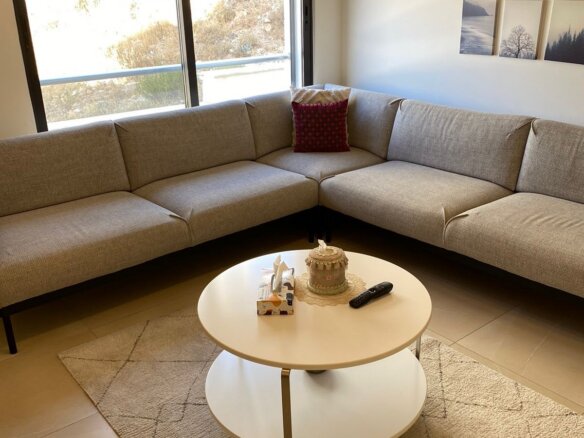 L-shaped beige sofa in a bright living room with a round white coffee table and a small cake, tissues, and a remote on top.