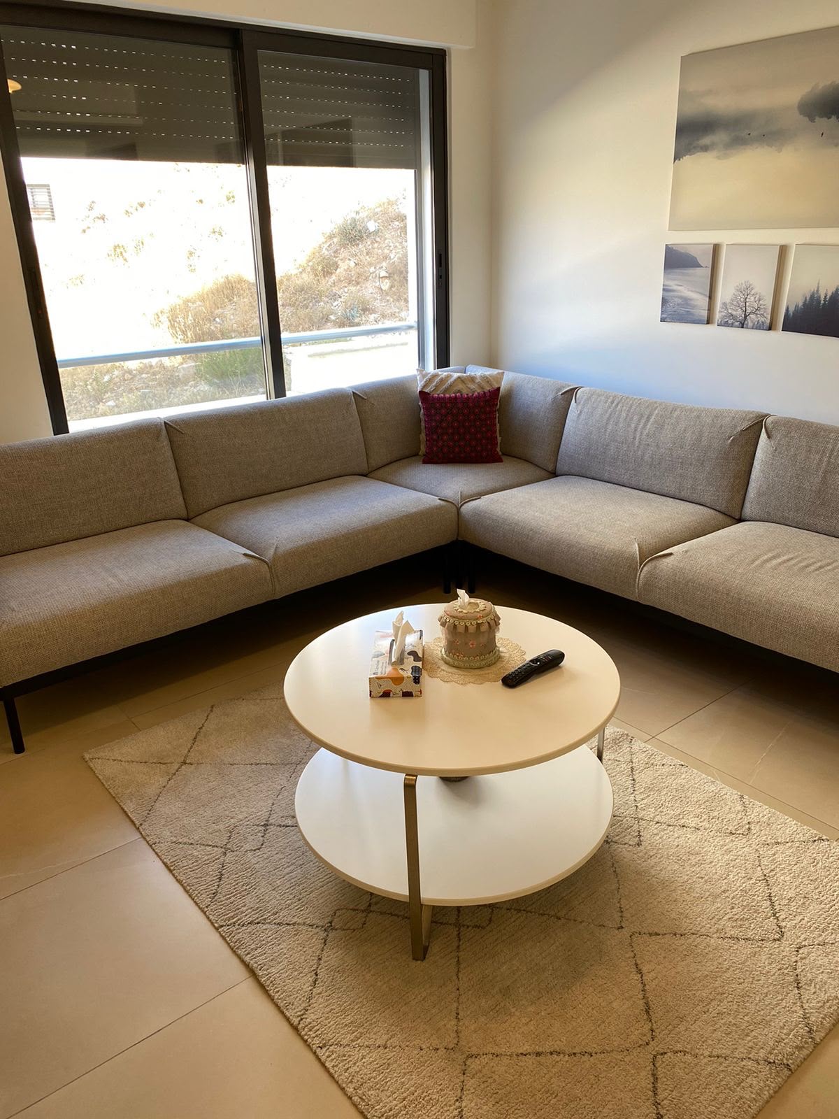 cec8705e-6530-4a53-92f5-d43cf0d9aeaf L-shaped beige sofa in a bright living room with a round white coffee table and a small cake, tissues, and a remote on top.